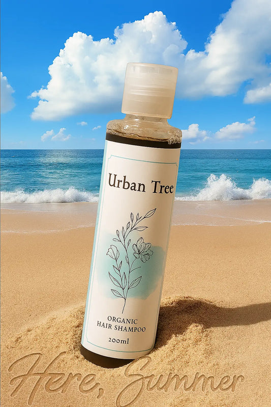 Organic Shampoo by Urban Tree placed on sandy beach with ocean waves and blue sky — summer-themed photo highlighting natural, refreshing hair care.