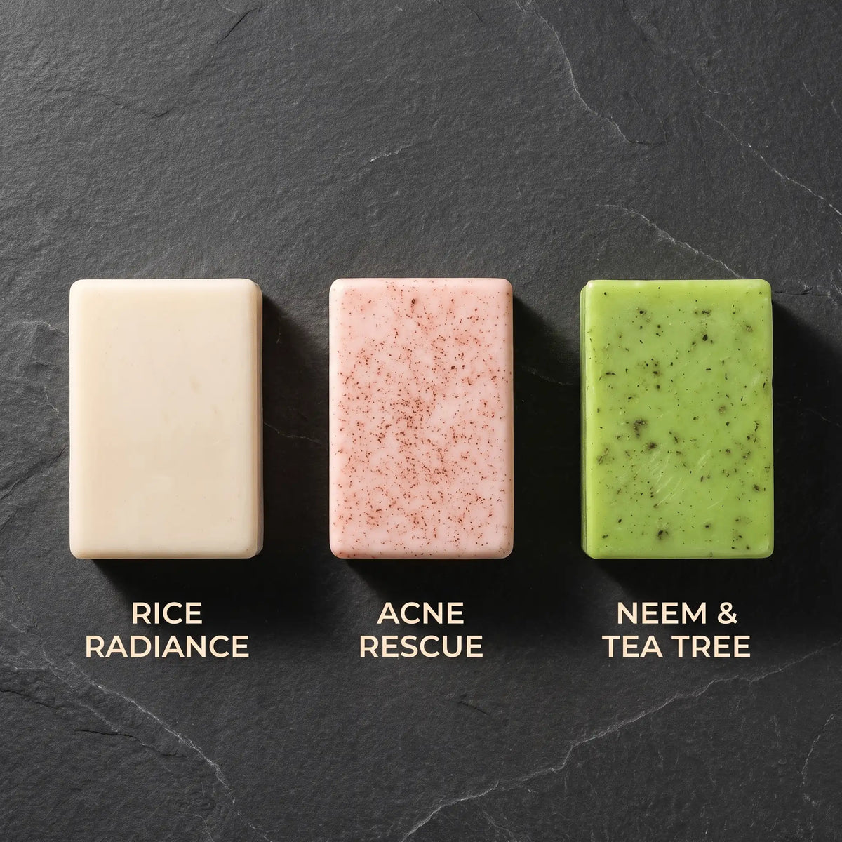 Overhead view of three Urban Tree goat milk soaps on dark slate, labelled Rice Radiance, Acne Rescue and Neem and Tea Tree